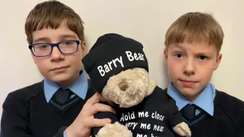 Schoolboys Oliver and Daniel hold up a stuffed bear. Both boys are in school uniform - Oliver is wearing glasses and has brown hair. Daniel has no glasses but fairer hair. He is raising an eyebrow. The stuffed bear has a hat on saying Barry Bear, and a T-shirt saying 'Hold me close, Carry me near, With me here, You have nothing to fear'.