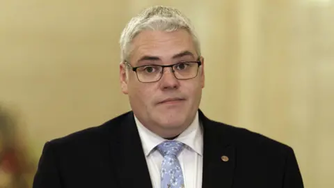 DUP leader Gavin Robinson, with short grey hair, wearing glasses, a white shirt, blue tie and black jacket. 