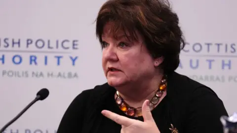 PA Media Fiona McQueen, who has short black hair, speaks into a microphone. She is wearing a black top and with her left index finger pointing to her right. 