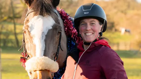 A woman wearing a black helmet and a dark red top, smiles as she stands next to a brown and white horse, which has blue and red tinsel around it.