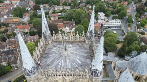 Joe Bilton/BBC Two men in harnesses with white helmets are standing at the top of a ladder looking at a golden weather vane at the top of a cathedral's pinnacle. The city of Lincoln is below them and in the distance.