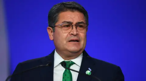 Honduras President Juan Orlando Hernández speaks in front of a blue backdrop. He has short hair and wears square-framed eye glasses. 