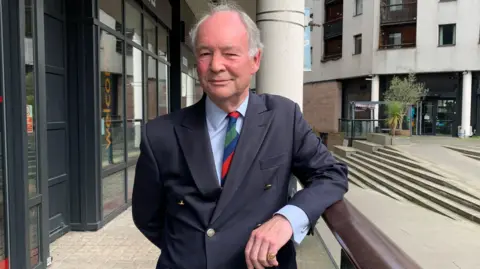 Philip Seccombe, the current Police and Crime Commissioner for Warwickshire Police. He is wearing a suit and tie and has his arm reesting on a balustrade.