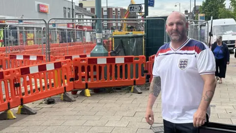 Roy Barrett a bald man with a white blue and red England shirt on and tattoos on his arms. He is stood outside his pub with the roadworks visible just behind him.