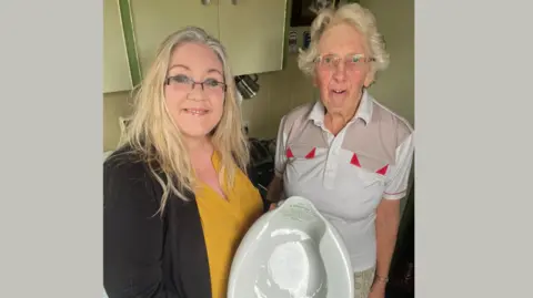 Julie Stevenson, in a yellow top and black cardigan is pictured holding a bedpan she has bought from Mary Jacobs, who stands to her right wearing a white shirt with red details.