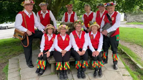 Four members of Furness Morris Dancers sat on a bench with six more standing behind them. They are all wearing black trousers, white shirts and red waistcoats. All but one have hats with flowers at the top. They are smiling. The phot was taken in 2023.