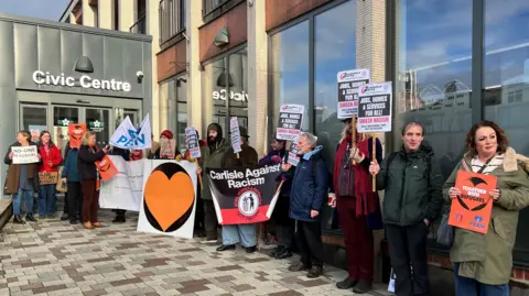 Just over a dozen people are standing outside the grey and cream civic centre building, holding various placards and signs. A large one reads "Carlisle against racism," and another says "No-one is illegal."