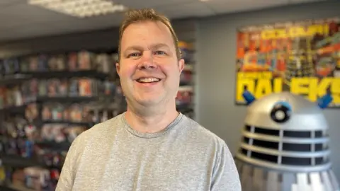 BBC / Jake Zuckerman Shopkeeper Paul Berry. He has short brown hair and is wearing a grey T-shirt. He is standing inside his shop with items on shelves and a large Dalek behind him