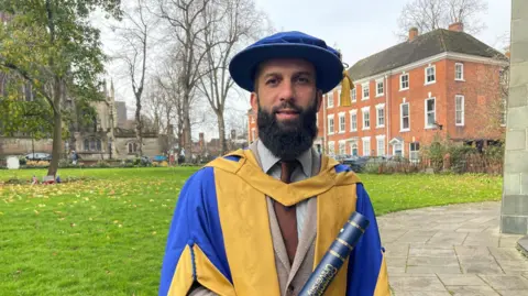 Moeen Ali wearing a yellow and blue robe holding a scroll from Coventry university