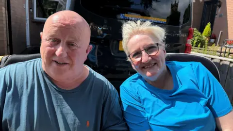 From left, John and Glenda O'Brien smiling as they look directly into the camera. They are sitting next to each other, outside the driveway of their home. John has a bald head and is dark wearing a blue T-shirt. Glenda has blonde hair and is wearing black glasses and a lighter blue T-shirt. A black van is parked behind them.