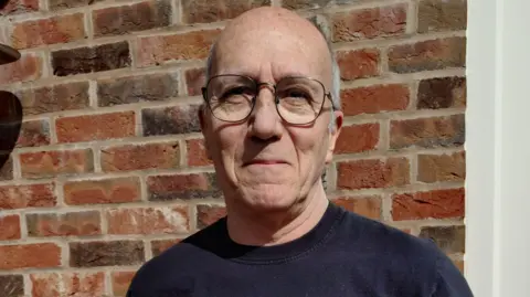 A man in a dark jumper and glasses stood in front of a brick wall. 