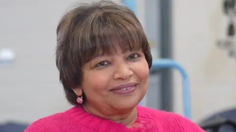 Jamie Niblock/BBC Prema Fairburn-Dorai smiles at the camera. She has a brown hair that is cut into a pixie bob with a fringe. She is wearing a pink jumper and has pink earrings in.