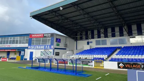 Ian Lamont Coleraine FC pitch. The seats in the stands are blue and white with an overhead cover. The blue dugout is at the sidelines of the pitch. A building with signs for 'Donaghy Bros' and 'Coleraine FC' on it is on the left. 