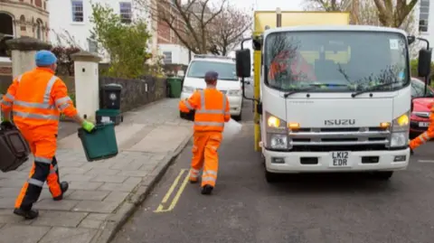 Refuse collectors wearing hi-vis orange suits on a residential road putting recycling into a refuse truck.