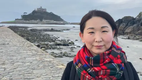 A woman stands on the beach in Marazion, St Michael's Mount is in the background.  She is wearing a tartan scalf and dark overcoat. 