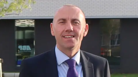 A head and shoulders shot of a man with a shaved head standing outdoors, smiling. He is wearing a dark suit jacket, a light blue checked shirt and a purple spotty tie. Behind him is a modern brick building with large reflective windows.