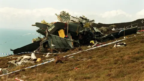 A photograph of the wreck of the Chinook helicopter, which had crashed in Scotland. It is on a large mound of grass, with police tape placed around the crash site.