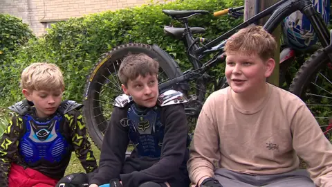 Three teenage boys sit in front of bikes leaning against a telegraph pole in front of a hedge. They are wearing protective body armour worn by cyclists. 