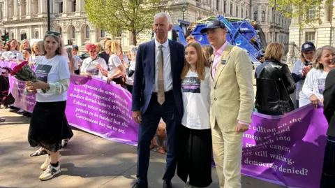 Chrissie Reidy/BBC A man in a blue suit, a woman in a white t-shirt and black dress and a man in a beige suit, is standing in front of a purple banner. There are a line of people in white t-shirts standing behind the banner.