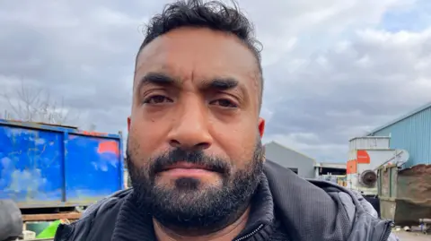 A man with black hair and a beard is looking at the camera. The sky is prominent in the background behind his head.