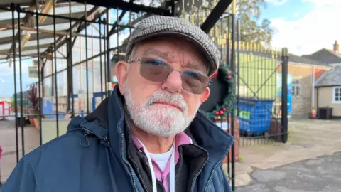 Man with glasses and flat cap stands in front of closed gate at Hythe ferry entrance