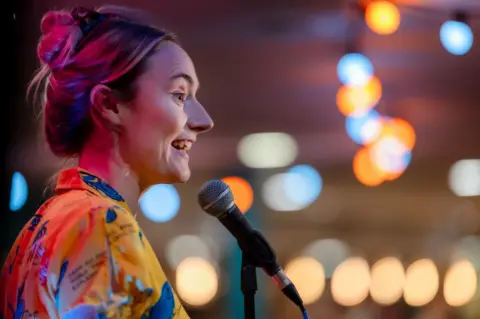 Tom Kay Steph Bradhsaw performing stand-up comedy. She is pictured side-on, speaking and smiling in front of a microphone. She has long blond hair pinned up in a messy bun. She is wearing an orange blouse with a blue motif.