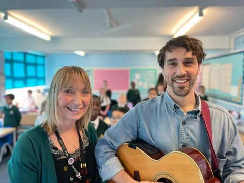 George Carden/BBC Two teachers standing in the foreground with one of them holding an acoustic guitar with children behind in the background across the classroom