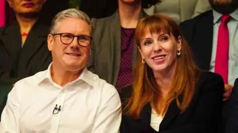 PA Media Starmer on the left in a white shirt with a mic pinned to it and Rayner next to him in a black blazer and white shirt. Both are sitting in an audience and smiling.