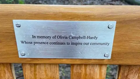 Yvonne Wright NEEDSPERMISS Memorial bench. Silver plaque reads: 'In memory of Olivia Campbell Hardy. Whose presence continues to inspire our community'