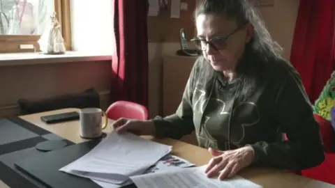 Jo Brearton is sat at desk looking at some papers spread out in front of her. She has long grey hair with part of it tied back. She is wearing glasses and a green jumper. 