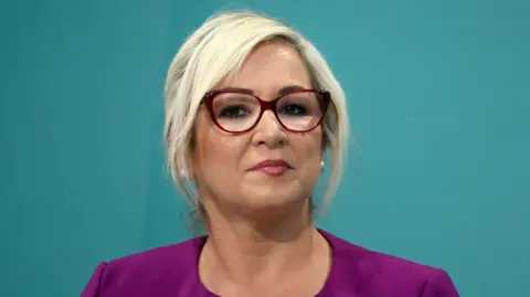 Getty Images Michelle O'Neill is wearing red glasses and pearl earrings. Her blond hair is tied back. A green backdrop is behind her. 
