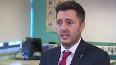 BBC Mr Morrison stands in a classroom with a white board and computer behind him. He has short black hair and beard and wears a navy suit with a white shirt and red tie.