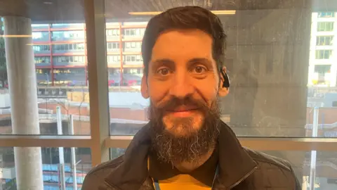 Gabriel Gabor, who has now found a job after previously sleeping rough on the streets of Manchester, smiles for the camera. He has a long brown-grey beard and wears a dark grey jacket and yellow jumper.