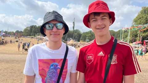 Richard Haugh/BBC Two boys side-by-side, both wearing bucket hats and bags over their shoulders. They are at a festival site, with people in the background.