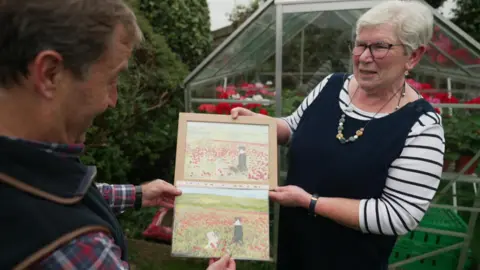 David Braine and Julie Taylor look at a framed painting featuring a black and white dog and a blond dog looking out at a filed of red poppies, and her photograph of the same scene.