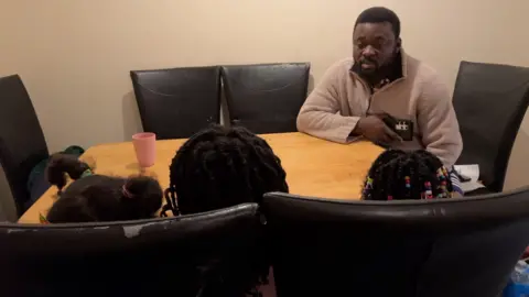 BBC Osei Kwaku sits at the table with his three primary school age daughters. Their backs are to the camera as their parents want to protect them from being identified.