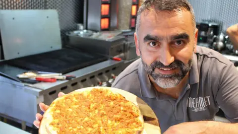 Mehmet Ulucan A smiling Mehmet Ulucan shows a plate of food to the camera in his restaurant kitchen. He has grey/black hair and a beard and wears a Mehmet Kitchen polo shirt.
