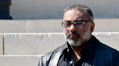 Nadeem standing on the steps of Cardiff Crown court. He has a white beard and grey short hard and rimmed glasses. 