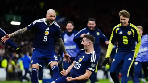 Several Scotland football players celebrate with delight after scoring a goal. One of the team slides on his knees across the turf while others race to hug him.