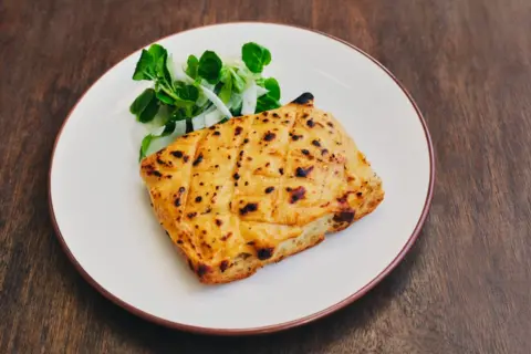 Michael Hani A round white plate sits on top of a brown, wooden, surface. On the plate is a yellow square of bread with melted cheese on top. To the side of the plate is a green leaf salad. 