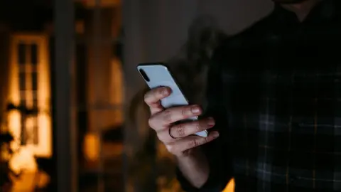 Getty Images An unidentifiable man in a dark room is shown holding a grey smartphone. He is wearing a check shirt.