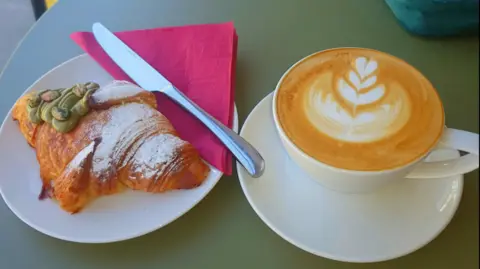 Zina A coffee and pistachio croissant at a cafe. They're both on white plates on a green table. The coffee has latte art on top.