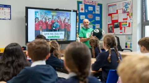 An NSPCC volunteer holds a mascot while talking to a class of primary school pupils during a Speak Out Stay Safe workshop. A presentation is up on the interactive whiteboard. The volunteer is wearing a green t-shirt.