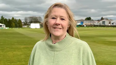 A woman in her late 50s is standing outside in the middle of a cricket field. She has blonde hair and is wearing a green jumper.