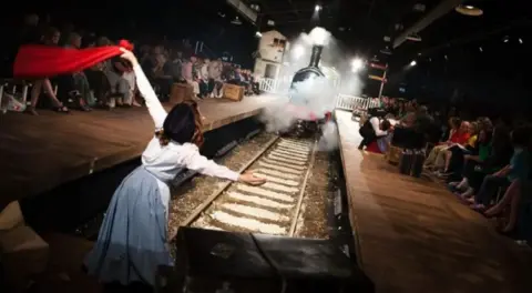 A picture of a steam train on a railway line in a specially adapted theatre with a woman stood in front of it waving a red cloth and audience members sat either side.