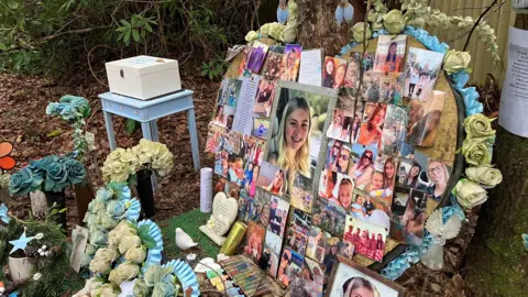 Picture shows a tree with a large heart - a metre tall and wide - made out of wood covered in pictures of Laionie from birth to teens. There are flowers in front, candles and ornaments.