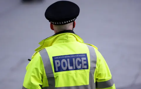 A police officer has his back to the camera. He is wearing a black police hat and is wearing a high-vis jacket with the words 'Greater Manchester Police' on the back.