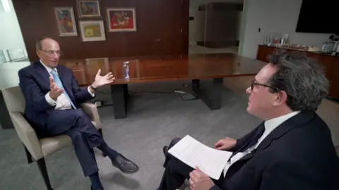 Wide shot showing Blackrock CEO Larry Fink being interviewed by BBC business editor Simon Jack. The two men are sitting across from each other with a long wooden table behind them and four pieces of art hanging on the wall