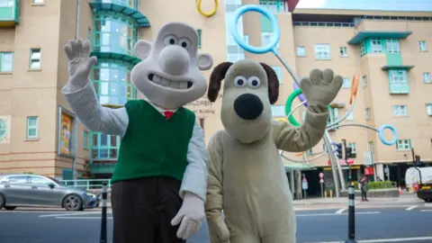 UHBW Two people dressed as Wallace and Gromit standing waving to camera outside the Bristol Children's Hospital, with the striking sculpture of huge multicoloured rings visible in the background