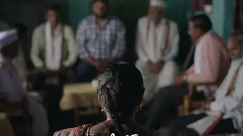 Courtesy: A view of Sthal Sthal: A match shows behind a woman in the foreground. She is facing a group of men who have come to assess her for the wedding match.